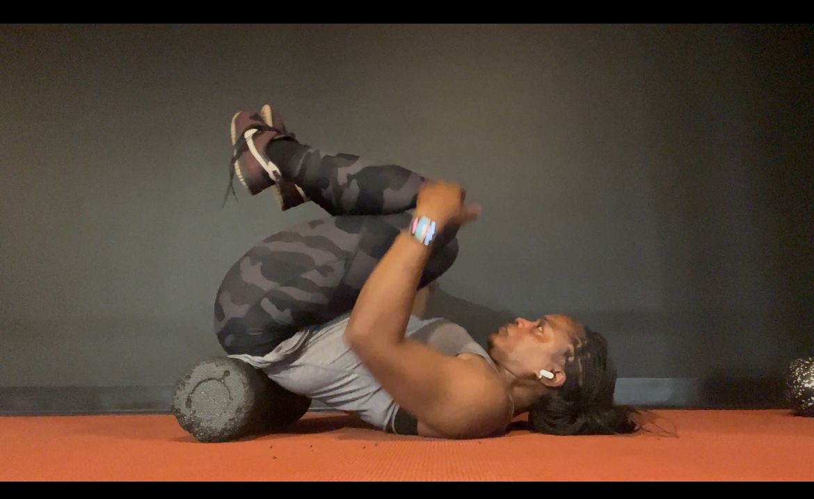 Person in gym clothes using a foam roller for back exercise, lying on an orange mat.